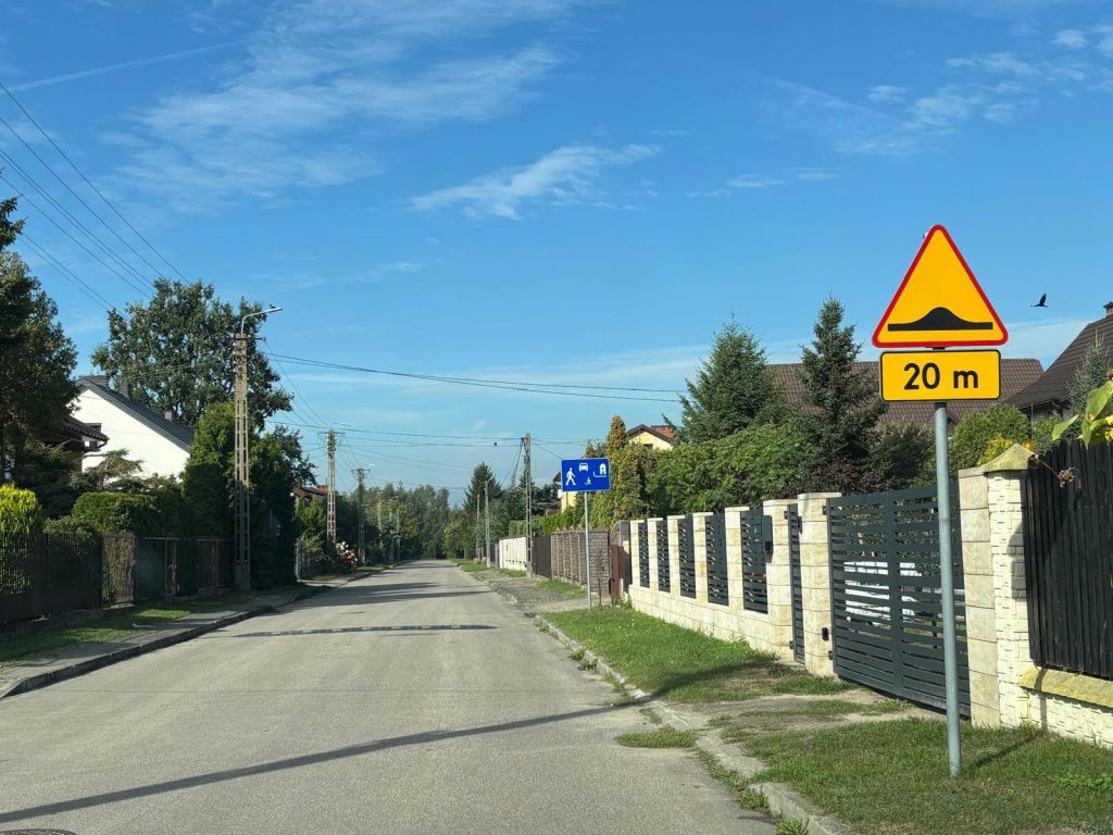 Droga asfaltowa z progiem zwalniającym, znak ostrzega 20 m przed nim. Zielone drzewa i krzewy wzdłuż ulicy. Niebieskie niebo z białymi chmurami w tle.