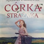 Kobieta w czerwono-białej sukience przy rowerze na tle zielonych pól. Napis Córka strażaka. Jasne, niebieskie niebo.