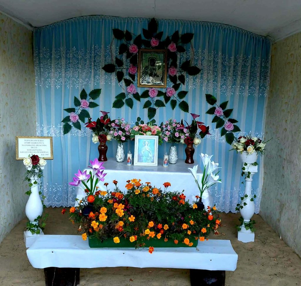 Ołtarz z niebieską zasłoną z kwiatami i obrazem Maryi. Wazon z kolorowymi kwiatami, w tym liliami i nagietkami. Tło zdobią różowe róże.