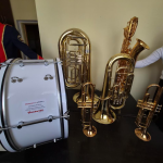 Na zdjęciu widać instrumenty muzyczne - tuby i bęben stoją na ciemnym stole. Złote tuby kontrastują z jasnym bębnem. Tło jest beżowe, a obok widoczny jest fragment ciemnego ubrania.