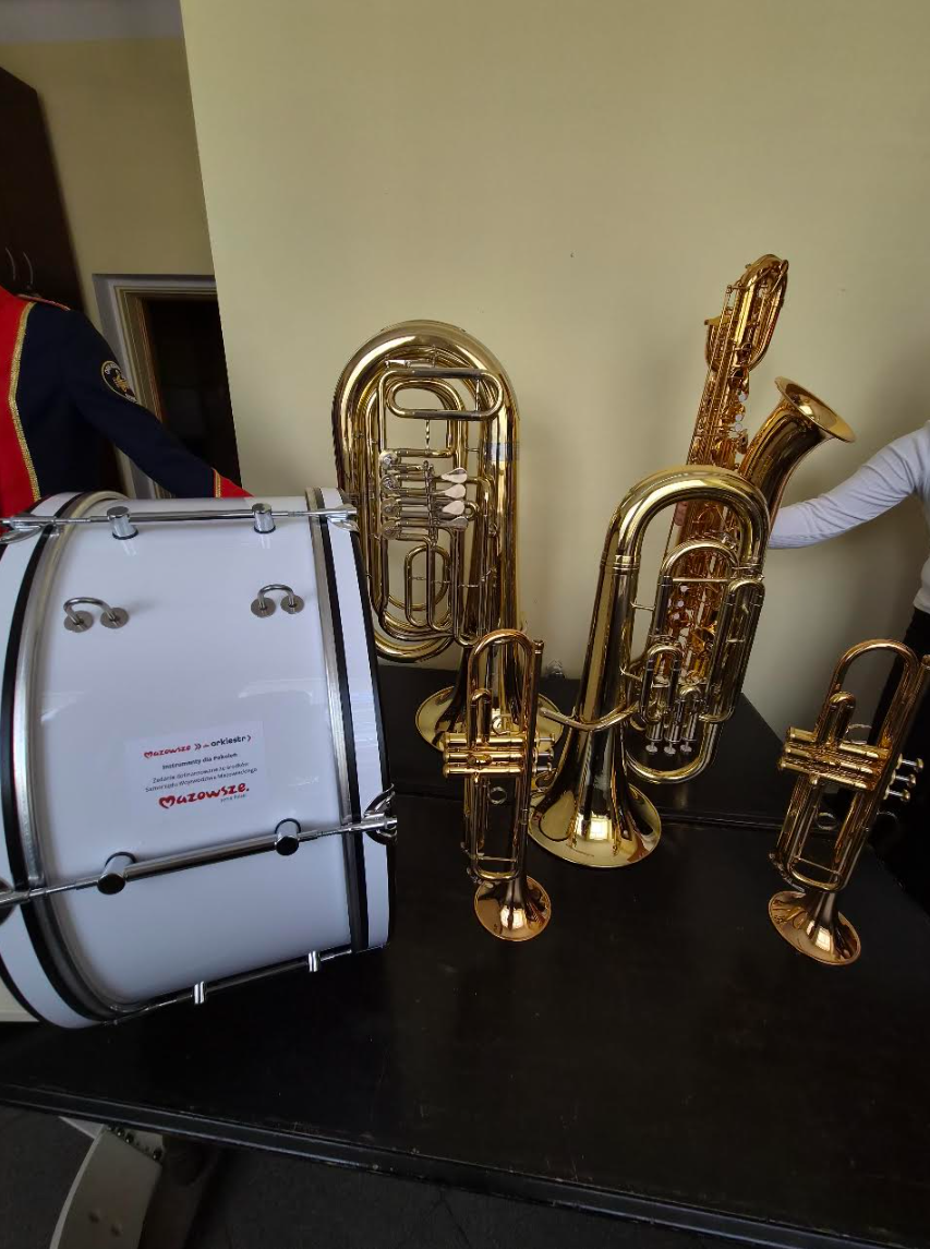 Na zdjęciu widać instrumenty muzyczne - tuby i bęben stoją na ciemnym stole. Złote tuby kontrastują z jasnym bębnem. Tło jest beżowe, a obok widoczny jest fragment ciemnego ubrania.