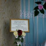 Tablica pamiątkowa w złotej ramie na tle niebieskiej zasłony z dekoracją kwiatową. Obok, białe i czerwone róże. Ściana w tle jest wzorzysta.