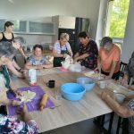xkr7eV1f Grupa osób przy dużym stole pracuje nad przygotowywaniem ciasta. Wnętrze jasnej kuchni z drewnianymi szafkami. Na stole różne akcesoria kuchenne i miski.