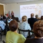 Na zdjęciu jest prezentacja z publicznością siedzącą w sali. Kobieta w fioletowym ubraniu stoi obok ekranu z slajdem. Tło jest jasne, widać biały sufit i kolorowe flagi.