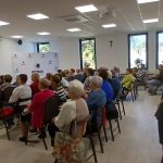 Grupa osób siedzi w jasnej sali na krzesłach, przy stole z naczyniami. W tle duże okna i biała ściana z logo. Sala ma biały sufit z kwadratowymi lampami.