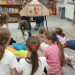 851981019_1262168322749790_8337163733784867813_n Dzieci siedzą na dywanie w bibliotece, oglądają teatrzyk. Kolorowe poduchy, półki z książkami w tle. Jasne wnętrze, dużo książek.