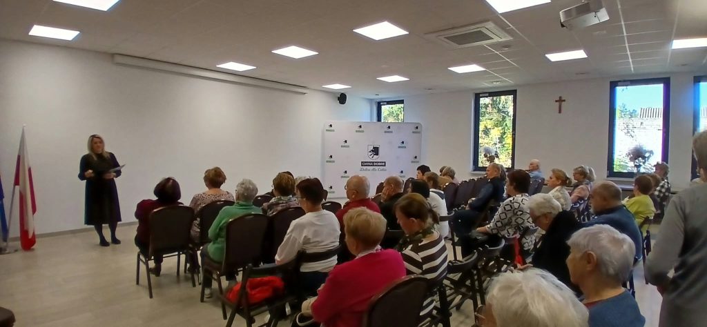 Sala z białymi ścianami, w której kobieta przemawia do grupy starszych osób. W tle biały baner z logiem Gmina Dobre. Na ścianach widoczne są okna i krzyż.