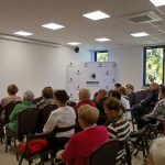 Sala z białymi ścianami, w której kobieta przemawia do grupy starszych osób. W tle biały baner z logiem Gmina Dobre. Na ścianach widoczne są okna i krzyż.