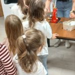 856271255_1896991514607059_3802146916130174800_n Osoby stoją w kolejce do stołu z urządzeniem. Tło to półki z książkami. Dominują kolory naturalne i czerwony.