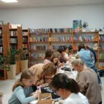 858353736_1134377599311355_4072352191280433879_n W pomieszczeniu z białym sufitem i regałami z książkami ludzie siedzą przy stołach, zajmując się rękodziełem. Tło wypełniają drewniane półki pełne kolorowych książek. Na stołach widoczne są przybory do malowania i kubki z napojami.