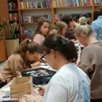 859272097_3676961245784297_3289728849357424418_n Osoby przy stole w bibliotece wykonują prace plastyczne. Półki z kolorowymi książkami w tle. Wnętrze jest jasne i przytulne.