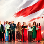 Grupa śpiewaków na scenie z tłem biało-czerwonej flagi. Mikrofony i nuty na boku. Kwiaty z przodu w biało-czerwonych barwach.