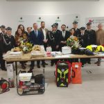 Grupa strażaków w galowych mundurach otacza stół z ekwipunkiem. Na stole są kaski, torby i bukiety kwiatów. Tło to jasna ściana z logo Powiat Miński.