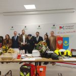 Grupa ludzi stoi za stołem z wyposażeniem strażackim. Tło to biały banner z logo i napisem Powiat Miński. Na stole leżą kwiaty, hełmy i torby.