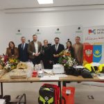 Na zdjęciu jest grupa osób stojących za stołem z zestawami ratunkowymi. Na stole leżą kwiaty i sprzęt w jaskrawych kolorach. W tle są banery z napisami i herbami.