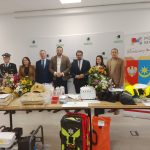 Grupa osób stoi przed stołem z wyposażeniem ratunkowym i kaskami. Tło zawiera logo Powiatu Mińskiego. Na stole są kwiaty i kartony.
