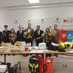 Na zdjęciu grupa ludzi stoi w pomieszczeniu obok stołu z hełmami i sprzętem ratowniczym. Osoby trzymają bukiety kwiatów, a w tle widoczny jest baner z herbem i napisem Powiat Miński. Kolory dominujące to jasne barwy, na tle biało-szaro-beżowego wnętrza.