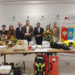 Grupa ludzi stoi za stołem pełnym sprzętu ratowniczego i kwiatów. Tło jest jasne z logo powiatu. Kaski i torby w jaskrawych kolorach.