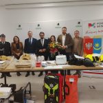 Na zdjęciu grupa osób stoi za stołem z sprzętem strażackim. Tło jest jasne, z logo powiatu i napisami. Obok widoczne są banery promocyjne i kwiaty.