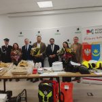 Grupa osób z kwiatami stoi przed stołem z hełmami i sprzętem ratowniczym. Tło ma białe ściany i bannery Powiatu Mińskiego. Kolory dominujące to czerń, biel i czerwień.