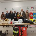 Grupa osób stoi przy stole pełnym sprzętu, w tle baner z logo powiatu. Dominują kolory jasne, kwiaty trzymane centralnie. Po lewej leży sprzęt ochronny i hełmy strażackie.