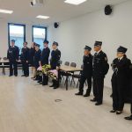 Grupa strażaków w galowych mundurach stoi w szeregu, trzymając bukiety kwiatów. Tło stanowi jasna sala z krzyżem i oknami. Podłoga jest drewniana, a obok znajdują się stoły i krzesła.