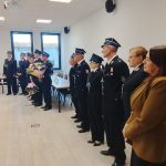 Grupa osób w jednolitych mundurach strażackich stoi w rzędzie, trzymając kwiaty. W tle jasne ściany i okna z widokiem na zewnątrz. Po prawej stronie stół z filiżankami.