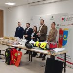 Grupa osób stoi przy stole z różnym sprzętem, na jasnym tle z logotypami. Na stole znajdują się hełmy, torby, generator. Obok stoi baner z herbem powiatu mińskiego.