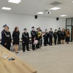 Grupa strażaków w mundurach stoi w szeregu w jasnym pomieszczeniu. Kilka osób trzyma bukiety kwiatów. W tle widać białe ściany i drewniane drzwi.