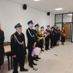 Grupa strażaków w eleganckich mundurach stoi w rzędzie z kwiatami. Tło jasne, z drewnianymi drzwiami i białą ścianą. Na podłodze jasne panele.