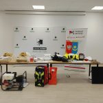 Stół z różnorodnym sprzętem strażackim na tle białych paneli z logami gminy i powiatu. Podkłady hełmów, torby ratunkowe, agregat. Utrzymanie w jasnych barwach, na jasnym tle.