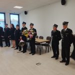 Czterech mężczyzn i trzy kobiety w mundurach strażackich stoi w jasnym pomieszczeniu z drewnianą podłogą i białymi ścianami. Dwie osoby trzymają kolorowe bukiety. W tle duże okna i stół.