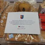 Zdjęcie przedstawia sprzęt OSP w workach na stole, z tabliczką informacyjną. Kolory dominujące: brązowy, biały, czerwony. W tle pudełka.