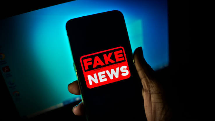 Osoba trzyma smartfon z wyświetlonym czerwono-białym napisem FAKE NEWS. Tło jest ciemne z niebieskawym monitorem.